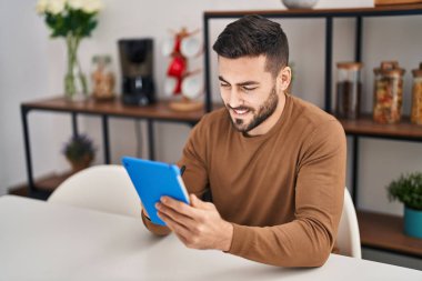 Young hispanic man using touchpad sitting on table at home