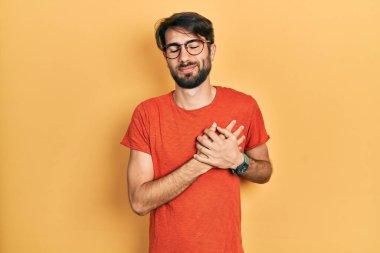 Gündelik kıyafetler ve gözlük takan genç İspanyol adam elleri göğsünde, gözleri kapalı ve yüzünde minnet dolu bir jest ile gülümsüyor. sağlık kavramı. 