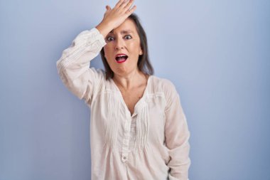 Middle age hispanic woman standing over blue background surprised with hand on head for mistake, remember error. forgot, bad memory concept. 