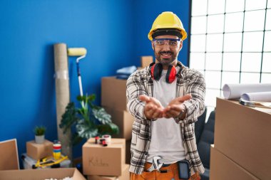 Evde çalışan sakallı, elleri avuçlarında gülümseyen ya da el hareketi yapan genç İspanyol bir adam. Bekleme ve koruma 