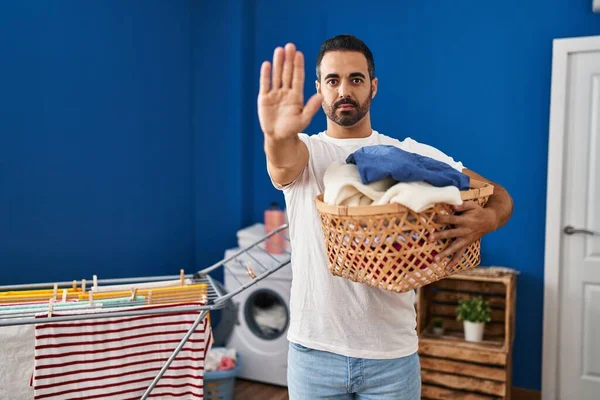 Young hispanic man with beard holding laundry basket with open hand doing stop sign with serious and confident expression, defense gesture
