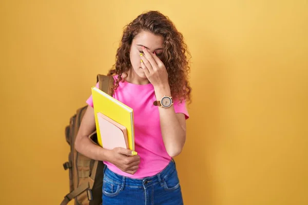Young caucasian woman wearing student backpack and holding books tired rubbing nose and eyes feeling fatigue and headache. stress and frustration concept.