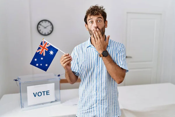 Young handsome man at political campaign election holding australia flag covering mouth with hand, shocked and afraid for mistake. surprised expression