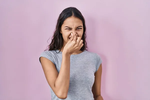 若いブラジル人女性はピンクの背景の上にカジュアルなTシャツを着て何か臭いと嫌な 耐え難い匂いを嗅ぎ 鼻の上に指で息を保持します 臭いが悪い — ストック写真