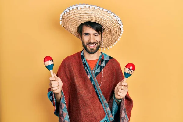 Young hispanic man wearing mexican hat holding maracas winking looking at the camera with sexy expression, cheerful and happy face.