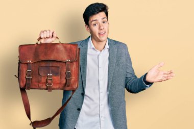 Young hispanic man holding business bag celebrating achievement with happy smile and winner expression with raised hand 