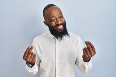 Mavi arka planda duran Afrikalı Amerikalı adam elleriyle para hareketi yapıyor, maaşını istiyor, milyonerlik işi yapıyor. 