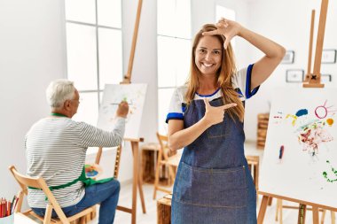 Hispanic woman wearing apron at art studio smiling making frame with hands and fingers with happy face. creativity and photography concept. 