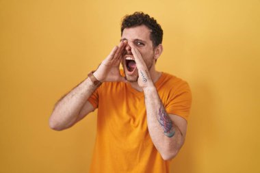 Sarı arka planda dikilen Latin asıllı genç adam elleri ağzında kızgın bir şekilde bağırıyor. 
