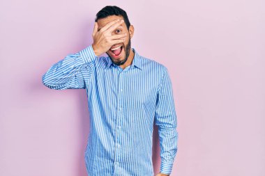 Hispanic man with beard wearing casual blue shirt peeking in shock covering face and eyes with hand, looking through fingers with embarrassed expression. 