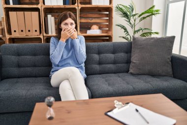 Young brunette woman at consultation office shocked covering mouth with hands for mistake. secret concept. 
