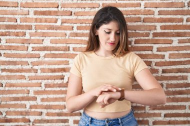 Genç esmer kadın tuğla duvarın üzerinde duruyor. Kol saatini kontrol ediyor. Rahat ve kendinden emin. 