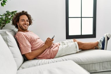 Genç İspanyol adam mutlu bir şekilde gülümsüyor. Evdeki kanepede oturmuş akıllı telefon kullanıyor..