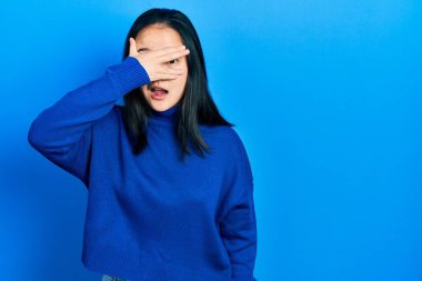 Young chinese girl wearing casual clothes peeking in shock covering face and eyes with hand, looking through fingers with embarrassed expression. 