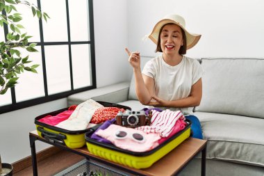 Beautiful middle age hispanic woman packing summer clothes in suitcase with a big smile on face, pointing with hand and finger to the side looking at the camera. 