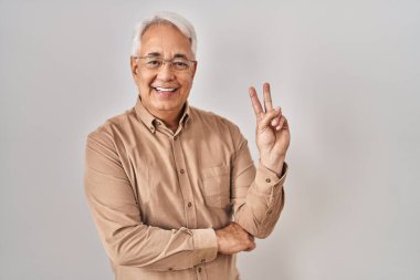 Hispanic senior man wearing glasses smiling with happy face winking at the camera doing victory sign. number two. 