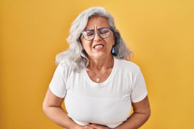 Middle age woman with grey hair standing over yellow background with hand on stomach because indigestion, painful illness feeling unwell. ache concept. 