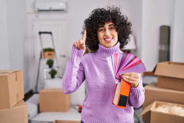 Young brunette woman with curly hair choosing color of new house wall surprised with an idea or question pointing finger with happy face, number one 