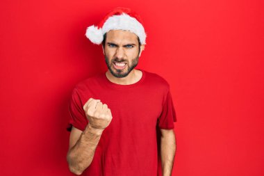 Young hispanic man wearing christmas hat angry and mad raising fist frustrated and furious while shouting with anger. rage and aggressive concept. 