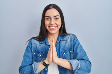 Hispanic woman standing over blue background praying with hands together asking for forgiveness smiling confident. 