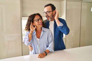 Middle age couple standing together smiling with hand over ear listening an hearing to rumor or gossip. deafness concept. 