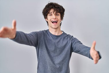 Young man standing over isolated background looking at the camera smiling with open arms for hug. cheerful expression embracing happiness. 