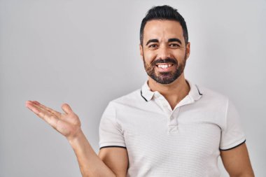 Young hispanic man with beard wearing casual clothes over white background smiling cheerful presenting and pointing with palm of hand looking at the camera. 