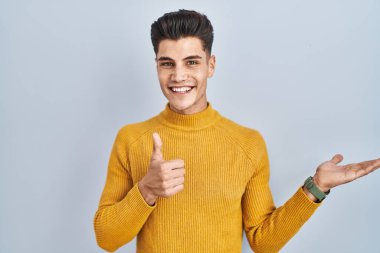 Young hispanic man standing over blue background showing palm hand and doing ok gesture with thumbs up, smiling happy and cheerful 