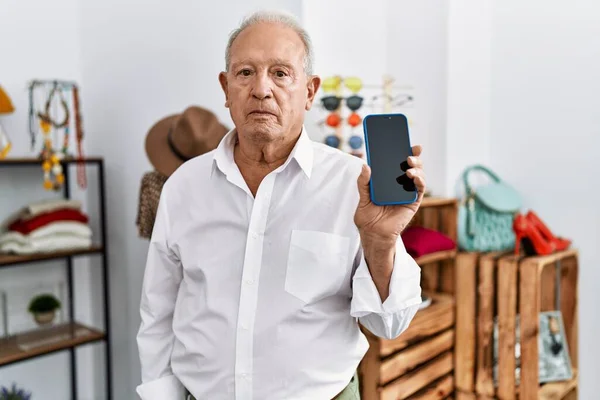 Senior man holding smartphone at retail shop thinking attitude and sober expression looking self confident