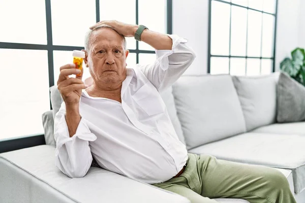 Senior man holding pills confuse and wondering about question. uncertain with doubt, thinking with hand on head. pensive concept.