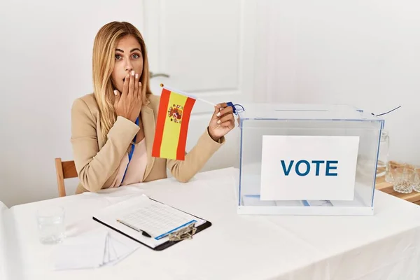 Blonde beautiful young woman at political campaign election holding spain flag covering mouth with hand, shocked and afraid for mistake. surprised expression