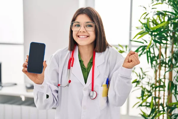 Young hispanic doctor woman showing smartphone screen screaming proud, celebrating victory and success very excited with raised arms 