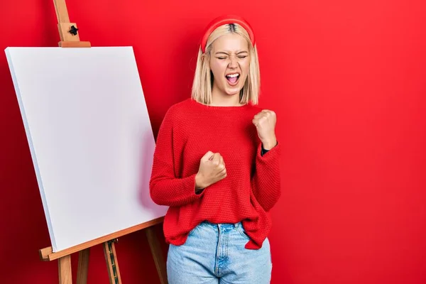 Beautiful Blonde Woman Standing Painter Easel Stand Celebrating Surprised Amazed — ストック写真