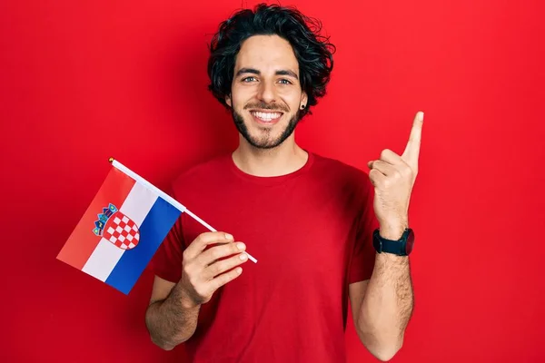 英俊的惊慌失措的男人举着克罗地亚国旗微笑着 带着一个想法或问题指指点点着幸福的脸 排名第一 — 图库照片
