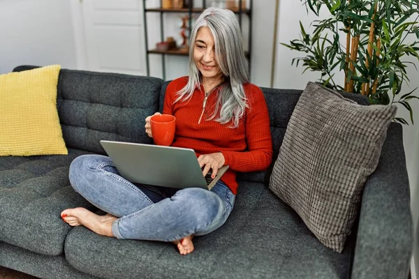 Middle age grey-haired woman drinking coffee using laptop at home