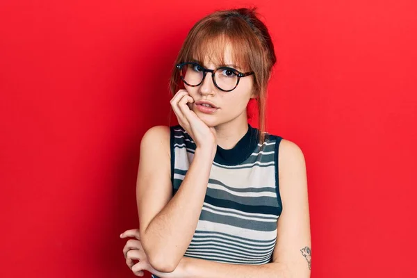 红头发的年轻女人穿着休闲装 戴着眼镜 双手放在咬指甲的嘴上 看起来很紧张 很紧张 焦虑问题 — 图库照片