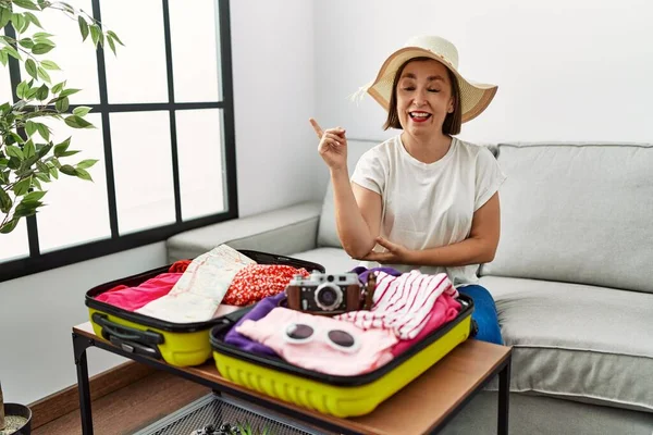 Beautiful middle age hispanic woman packing summer clothes in suitcase with a big smile on face, pointing with hand and finger to the side looking at the camera.