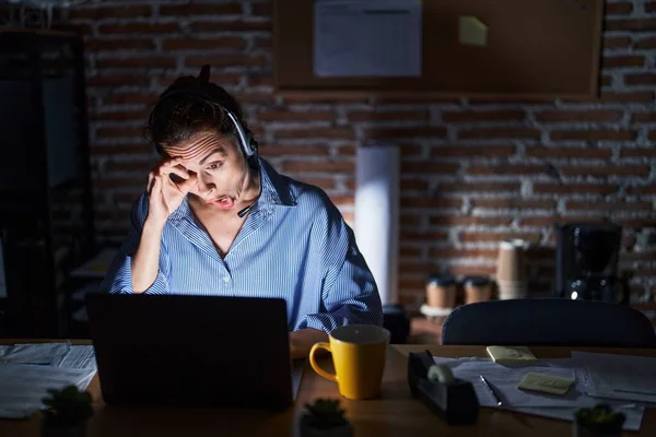 Beautiful brunette woman working at the office at night doing ok gesture shocked with surprised face, eye looking through fingers. unbelieving expression.