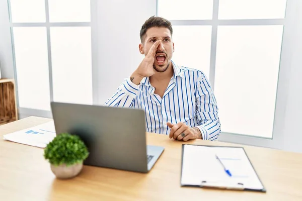 Young handsome man with beard working at the office using computer laptop shouting and screaming loud to side with hand on mouth. communication concept.