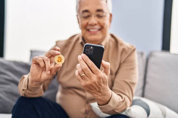 Senior man using smartphone holding bitcoin at home