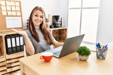 Young brunette woman working at the office with laptop smiling cheerful presenting and pointing with palm of hand looking at the camera. 