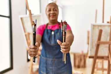 Kıdemli Afro-Amerikan kadın resim stüdyosunda elinde fırçayla gülümsüyor.