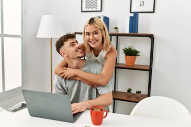 Genç, beyaz bir çift kucak açıp dizüstü bilgisayar kullanıyor. Evdeki masanın üstünde oturuyorlar..