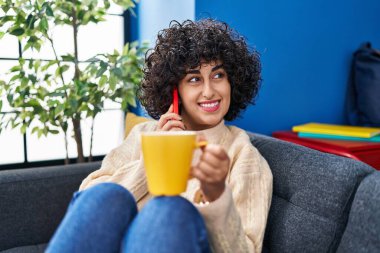 Orta Doğulu genç bir kadın kahve içiyor ve evde akıllı telefondan konuşuyor.