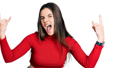 Young brunette teenager wearing red turtleneck sweater shouting with crazy expression doing rock symbol with hands up. music star. heavy music concept. 