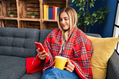 Genç sarışın kadın kahve içiyor ve evde akıllı telefon kullanıyor.