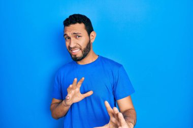 Hispanic man with beard wearing casual blue t shirt disgusted expression, displeased and fearful doing disgust face because aversion reaction. with hands raised 