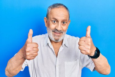 Handsome senior man with beard wearing casual white shirt approving doing positive gesture with hand, thumbs up smiling and happy for success. winner gesture. 