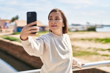 Orta yaşlı bir kadın, deniz kenarındaki akıllı telefonun yanında kendinden emin bir şekilde selfie çekiyor.