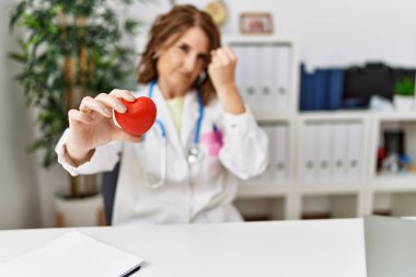 Klinikte kalbi tutan orta yaşlı bir doktor. Gülümsüyor, eli ve parmağı yan tarafa uzanıyor. 